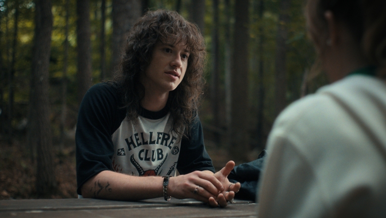 A young man with long, messy brown hair sitting at a picnic table in the woods; still from "Stranger Things 4."
