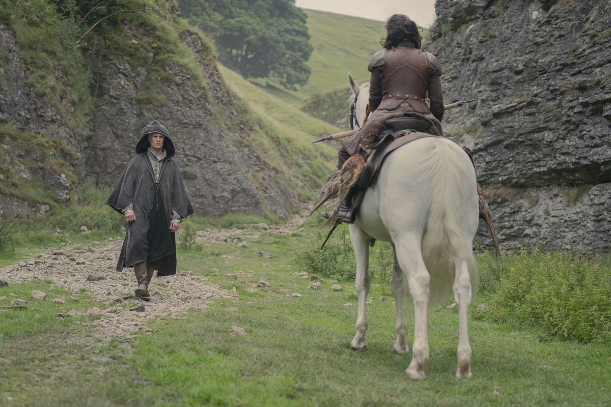 Daemon standing with a cloak and his hood up, walking towards a woman on the horse with her back turned to us